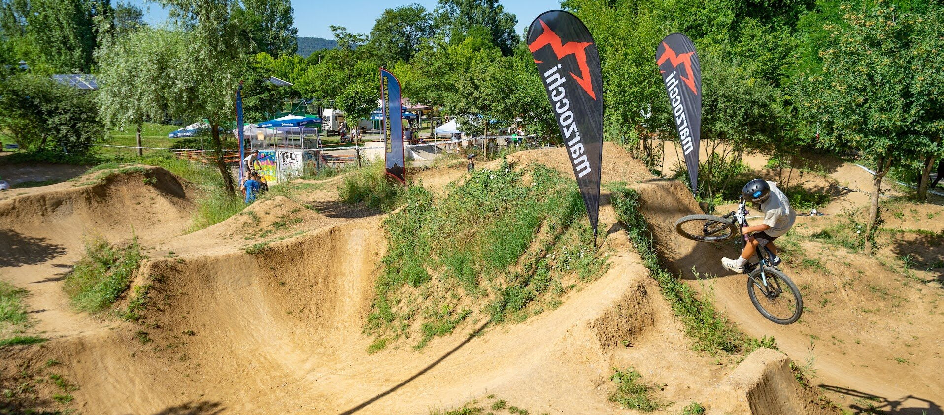 Ein Mountainbiker mit Helm fährt auf einem sandigen Pumptrack mit mehreren Kurven und Sprüngen. Zwei schwarze Fahnen mit rotem Logo und dem Schriftzug 'Marzocchi' stehen am Rand der Strecke. Im Hintergrund sind Bäume, Sonnenschirme und weitere …