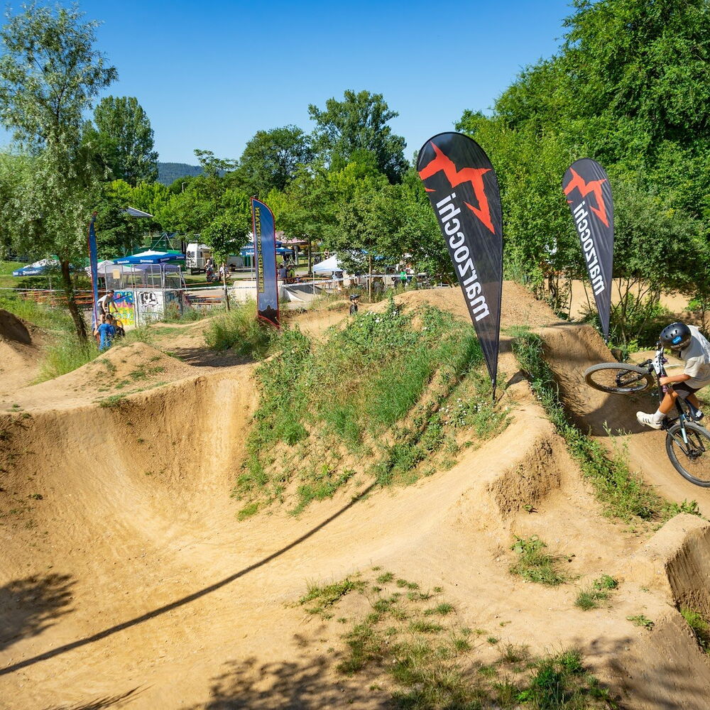 Ein Mountainbiker mit Helm fährt auf einem sandigen Pumptrack mit mehreren Kurven und Sprüngen. Zwei schwarze Fahnen mit rotem Logo und dem Schriftzug 'Marzocchi' stehen am Rand der Strecke. Im Hintergrund sind Bäume, Sonnenschirme und weitere …