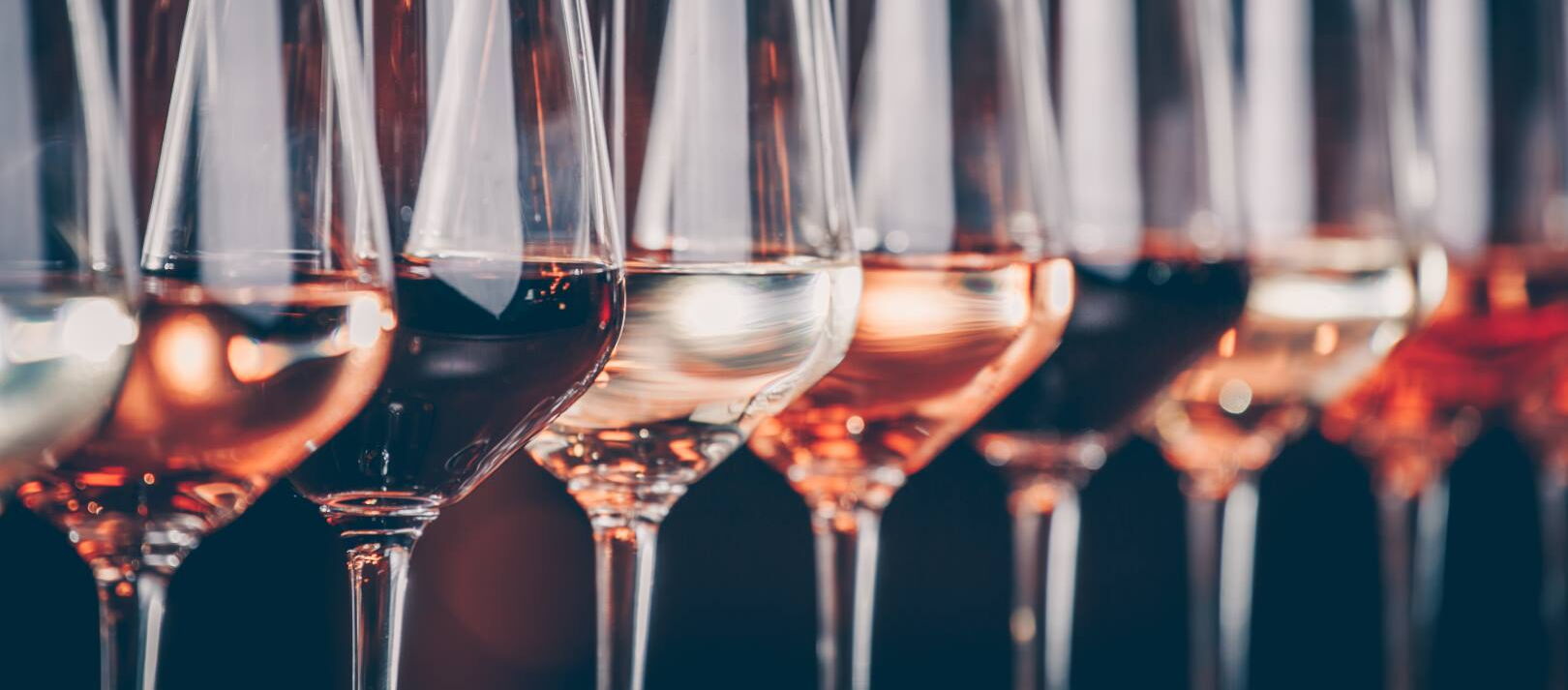 Mehrere Weingläser stehen nebeneinander, gefüllt mit Weißwein, Roséwein und Rotwein in unterschiedlichen Farbtönen. Die Gläser sind aufgereiht und spiegeln das Licht wider.