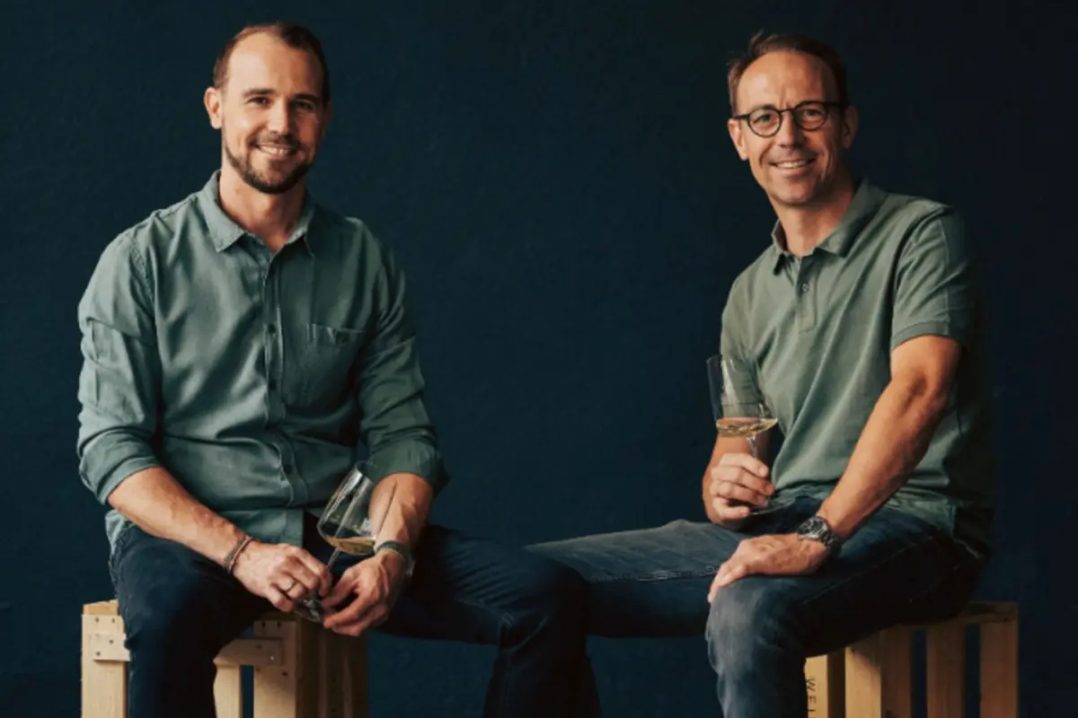 Zwei Männer sitzen auf Holzkisten vor dunklem Hintergrund und halten jeweils ein Glas Weißwein in der Hand. Beide tragen grüne Hemden und lächeln in die Kamera.