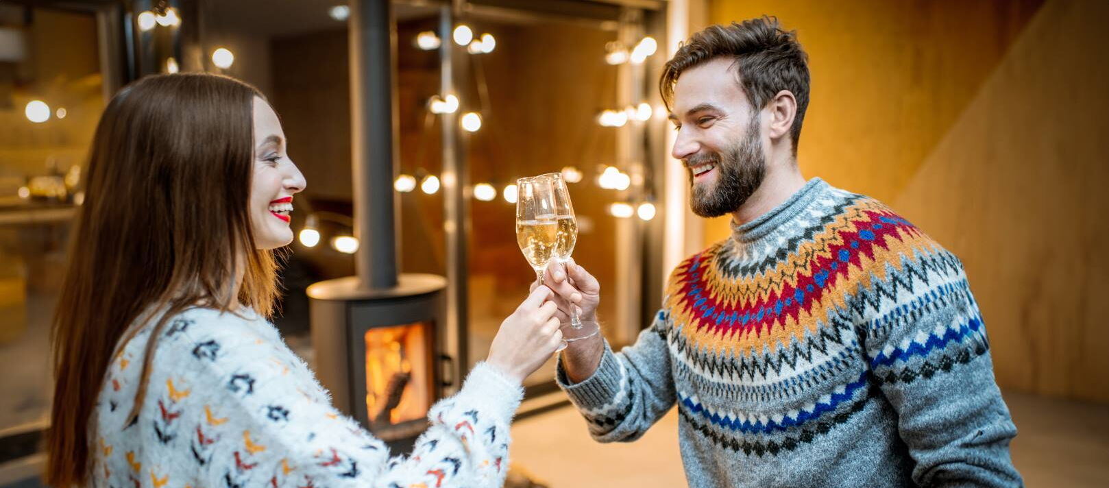 Eine Frau und ein Mann stehen sich gegenüber und stoßen mit gefüllten Sektgläsern an. Beide tragen bunte Strickpullover und lächeln sich an. Im Hintergrund brennt ein Kaminofen, warme Beleuchtung sorgt für gemütliche Atmosphäre.
