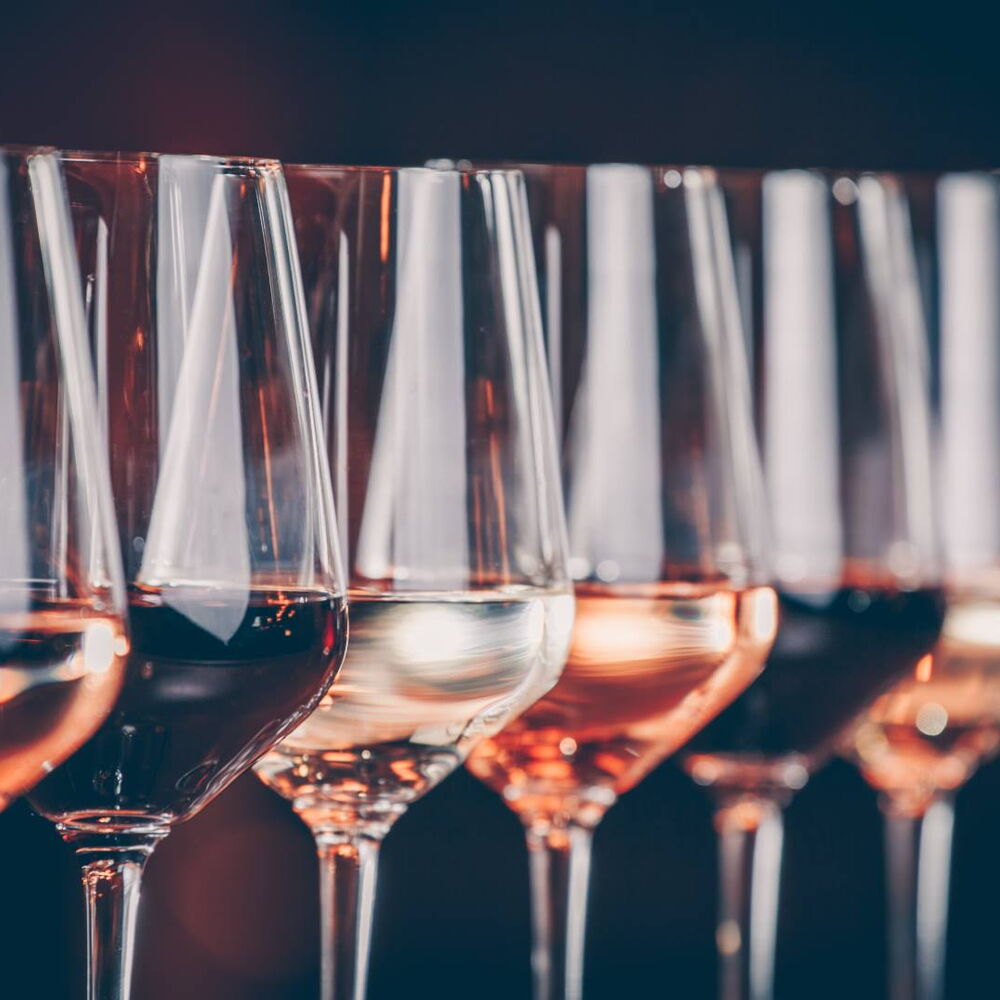 Mehrere Weingläser stehen nebeneinander, gefüllt mit Weißwein, Roséwein und Rotwein in unterschiedlichen Farbtönen. Die Gläser sind aufgereiht und spiegeln das Licht wider.