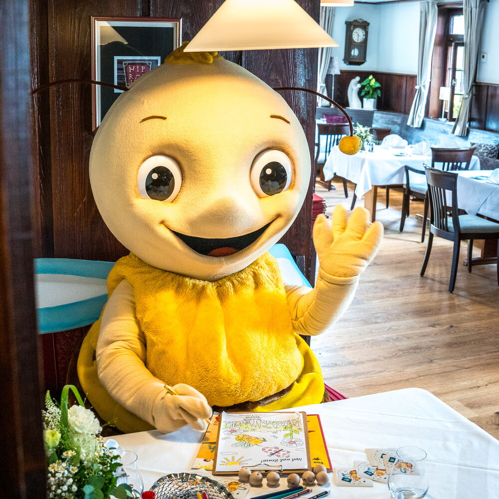 Großes Bienenmaskottchen mit gelbem Fell und Flügeln sitzt an einem gedeckten Tisch in einem Restaurant, winkt freundlich und hat Malutensilien vor sich liegen. Im Hintergrund sind weitere gedeckte Tische und Holzvertäfelung zu sehen.