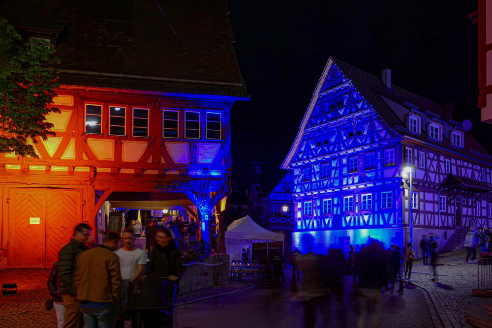 Zwei historische Fachwerkhäuser sind nachts farbig beleuchtet, das linke Haus in Rot-Orange, das rechte in Blau. Dazwischen stehen und gehen mehrere Menschen, einige sind unscharf in Bewegung. Im Hintergrund ist ein weißes Zelt aufgebaut, die Szene …