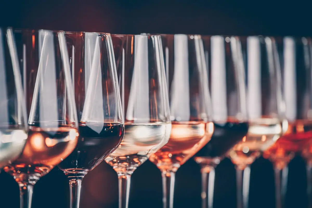 Mehrere Weingläser stehen nebeneinander, gefüllt mit Weißwein, Roséwein und Rotwein in unterschiedlichen Farbtönen. Die Gläser sind aufgereiht und spiegeln das Licht wider.